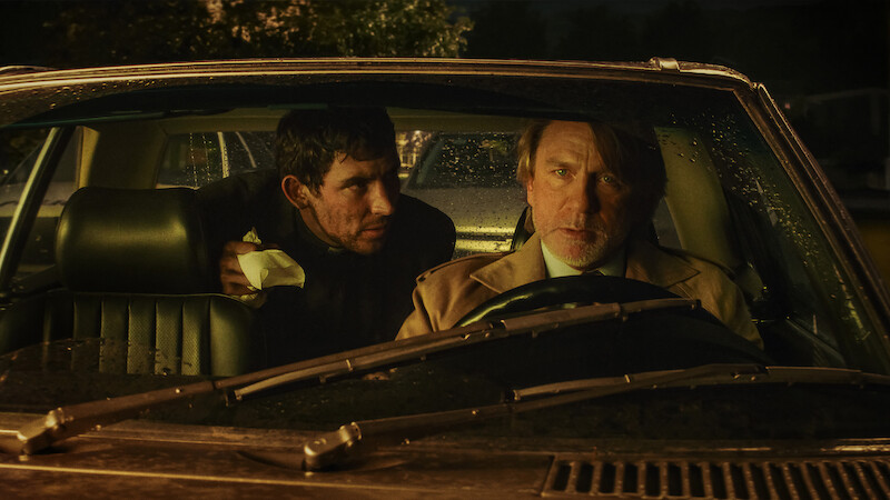 Two men sit in a car at night, illuminated by dim streetlights and rain on the windshield. The driver looks concerned while the passenger leans in with urgency, creating a tense and suspenseful atmosphere.