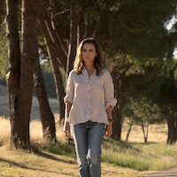 Woman with wavy hair in a light shirt and jeans walking outdoors on a path beside tall trees in a sunlit, grassy park or natural setting, looking serious.
