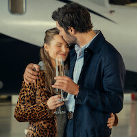 A couple embraces affectionately while holding champagne glasses in front of a private jet inside an aircraft hangar, suggesting a luxurious or celebratory moment.