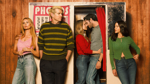 Five people in casual clothes pose in front of a vintage photo booth with wood-paneled walls; two people kiss inside the booth while the others stand or lean nearby, creating a retro and relaxed atmosphere.