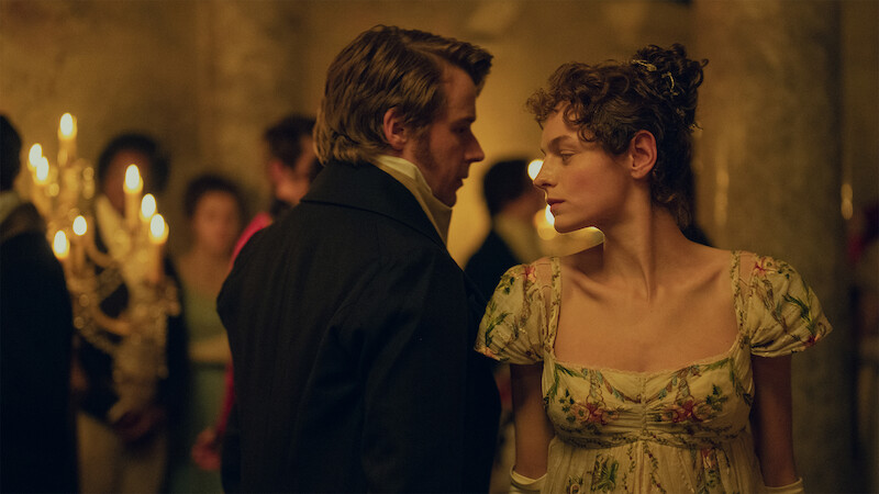 Two people dressed in historical costumes stand close together in an elegant ballroom, surrounded by softly lit chandeliers and guests, suggesting a formal or romantic setting from a past era.