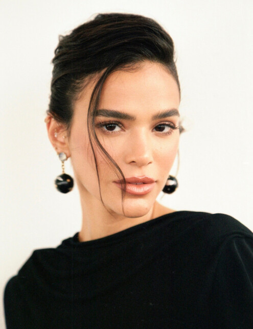Bruna Marquezine é uma mulher branca, magra, de cabelos escuros e presos, com brincos grandes e veste um vestido preto