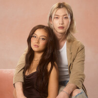 Two people pose together in a studio with a soft pink background; one sits on a pink couch wearing glasses and a dark shirt, while the other, with long blonde hair and a tan jacket, sits close behind, both looking at the camera.