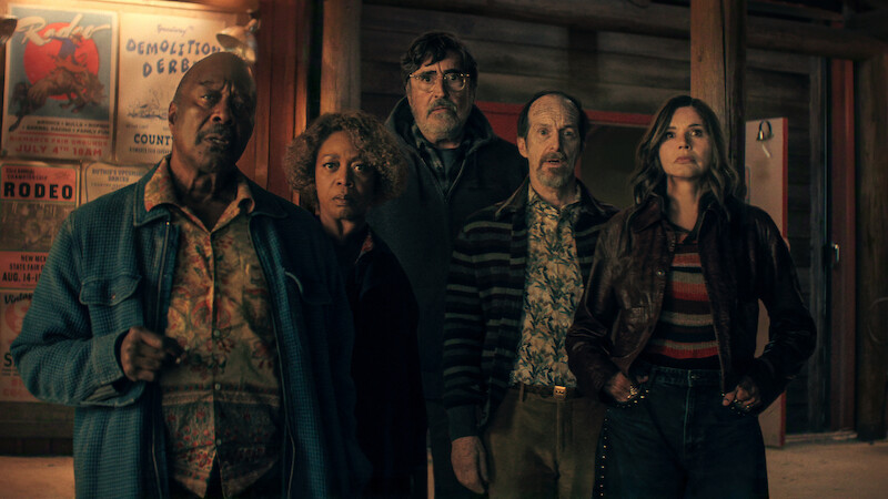Five people stand indoors in front of wooden walls with posters, looking intently ahead, wearing retro-style clothing in a dimly lit setting suggestive of a bar or lodge.