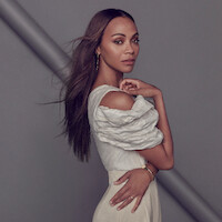 Zoe Saldaña stands against a gray background wearing an ensemble made of different shades of cream along with some gold jewelry, facing the camera with her hand touching her shoulder. 