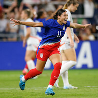 Soccer player in blue and red uniform celebrates on field with arms outstretched during a match, with blurred teammates and spectators in the stadium in the background.