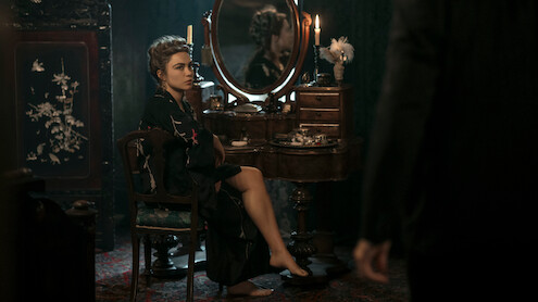 Woman in a dark robe sits at a vintage vanity with candles and flowers in a dim, elegant room, looking toward a standing figure partially visible in the foreground.