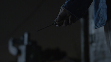 A person holding a knife in their right hand at night, wearing a dark blue jacket. The background is dimly lit, creating a suspenseful or tense atmosphere.