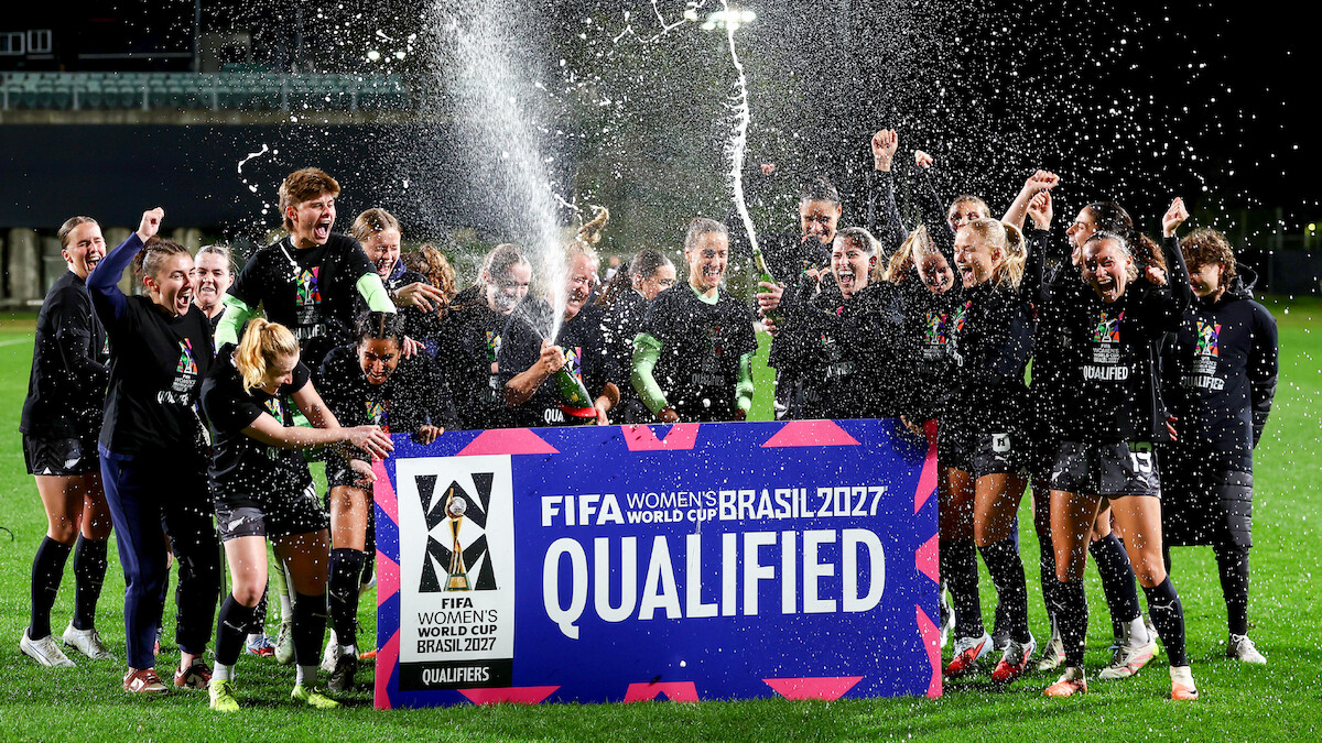 Women's soccer team celebrates at night on a field, spraying champagne and cheering behind a large sign reading "FIFA Women's World Cup Brasil 2027 Qualified." Bright stadium lights and festive atmosphere.