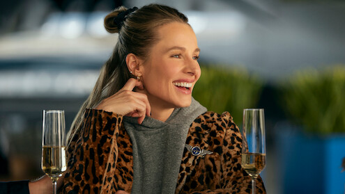 Smiling woman in leopard print coat sitting at a table with champagne glasses in a bright, modern setting, plants blurred in the background, appearing relaxed and joyful.