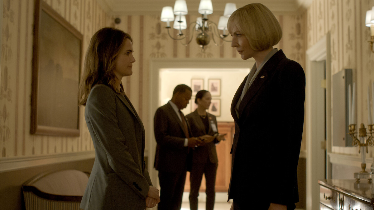 Two women in business suits face each other in a well-lit hallway with elegant decor; two other people are talking in the background, suggesting a formal or professional setting.