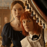 Two women in vintage clothing stand closely together, peering out from behind ornate curtains in an elegant, softly lit room with classic decor and floral arrangements, suggesting a historical or period drama setting.