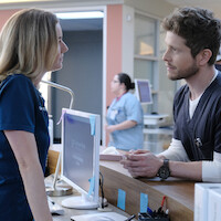 Two healthcare professionals in scrubs have a serious conversation at a hospital reception desk with computers, while another staff member works in the background.
