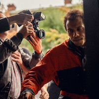 Man in red and black jacket pushing through crowd of photographers outdoors, with cameras pointed at him, surrounded by people and greenery in the background.
