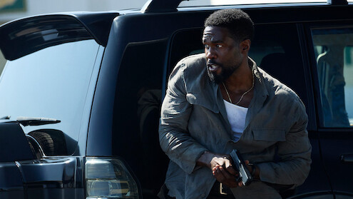 Man cautiously exits black SUV holding a handgun, wearing a gray jacket and white shirt, appearing alert in a tense urban outdoor setting.