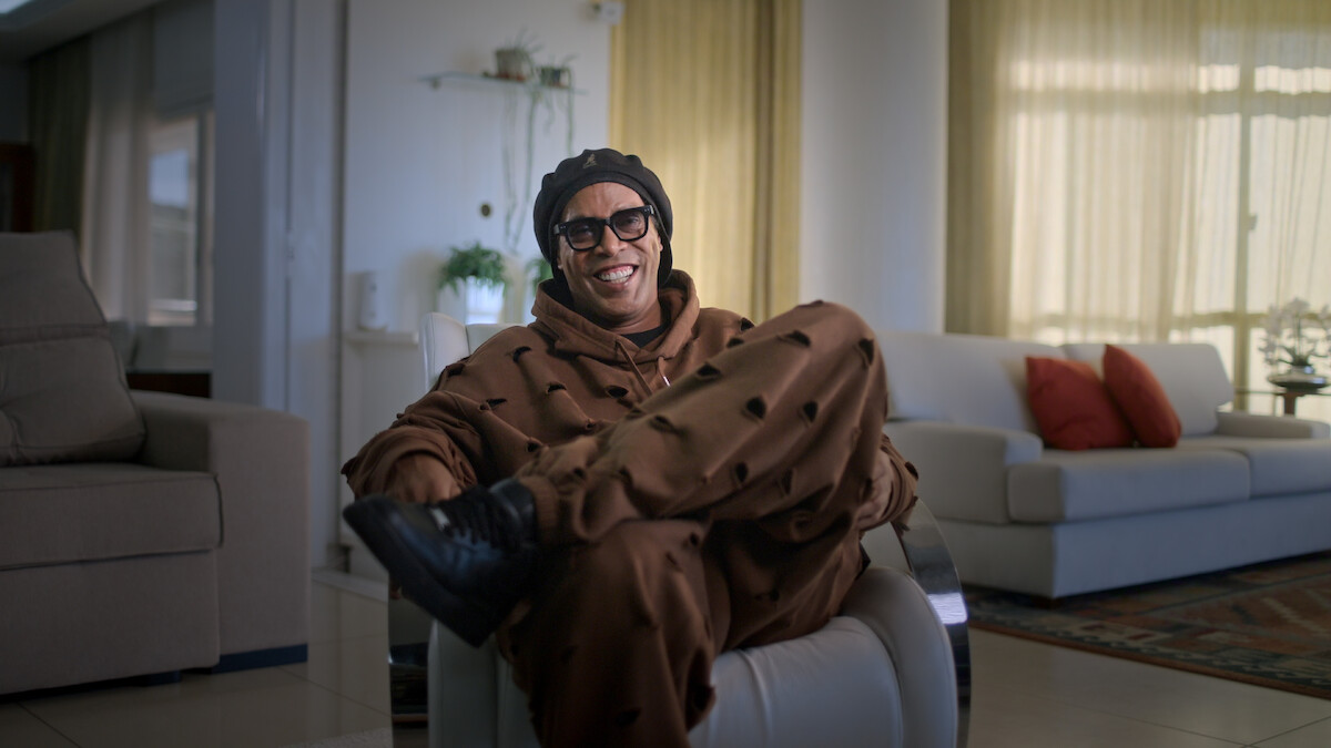 Ronaldinho Gaúcho in brown outfit and sunglasses smiles while sitting cross-legged on a modern chair in a bright living room with beige couches, red pillows, large windows, and natural light.