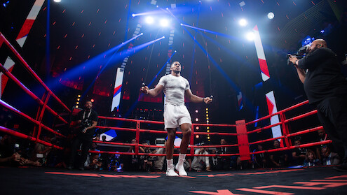 Boxer in a ring celebrates with a raised, gloved fist under dramatic arena lighting, surrounded by a cheering crowd, capturing a victorious and energetic mood.