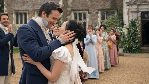 A bride and groom in period costumes embrace and smile outdoors, surrounded by cheering guests in Regency-era attire, with an old stone building and greenery in the background.
