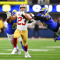 Football player in red and gold uniform runs with the ball on a stadium field, pursued by two defenders in blue and yellow uniforms during an NFL game.