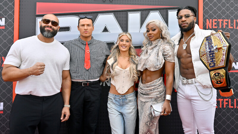 Rusev, Kit Wilson, Tiffany Stratton, Jade Cargill, and Trick Williams posing together on a red carpet at a WWE RAW event, with a branded backdrop. One person is holding an NXT Championship belt. The group is dressed in trendy and distinctive outfits.