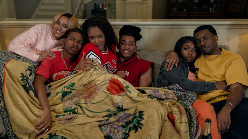 Six young adults sitting closely on a couch under a floral blanket in a cozy living room, smiling and cuddling, creating a warm and friendly atmosphere.