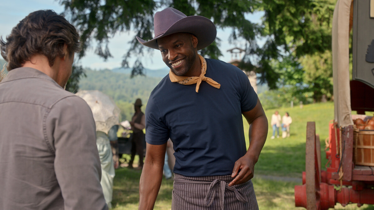 A smiling man in a cowboy hat and neckerchief stands outdoors near a wooden wagon, talking to another person. The setting is a green park or field with trees and people in the background, suggesting a friendly, rustic event.