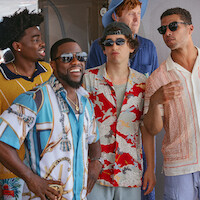 Five men wearing colorful shirts and sunglasses stand together outdoors, laughing and talking in a casual, sunny setting.