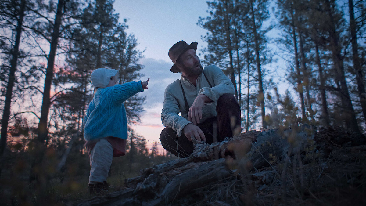 A man in a hat and a child in a blue sweater explore a forest at dusk, surrounded by tall trees. The child points at something, creating a sense of curiosity and peacefulness in the natural setting.