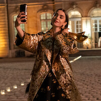 A woman in a luxurious gold-patterned coat takes a selfie while holding gold high heels outside an elegant, illuminated building at night.