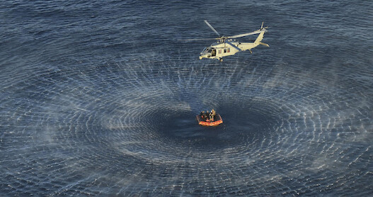 A helicopter hovers above the ocean, creating circular waves while performing a rescue operation near a life raft with several people aboard.