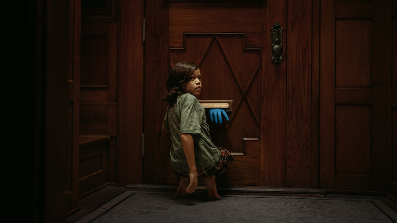 A young boy sits in front of a door as a blue glove reaches through the mail slot.