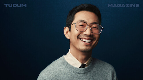 Lee Sung Jin stands in front of a backdrop that shifts from black to dark blue, smiling and looking to camera.