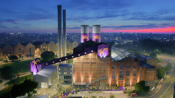Large whimsical chocolate factory with purple “W” logos, brick exterior, and tall chimneys, set in a city at dusk. Brightly lit, surrounded by greenery and roads, with apartments in the background under a colorful sky.