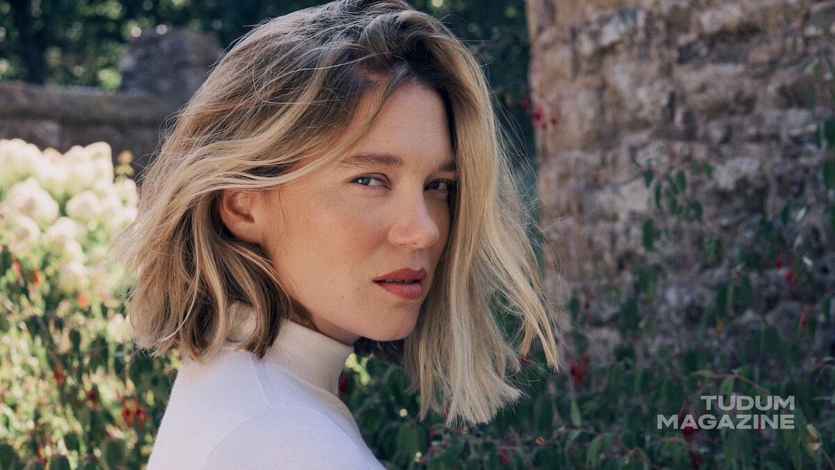 A woman with shoulder-length blonde hair in a white top stands outdoors near a stone wall and green foliage, with sunlight illuminating her face and flowers in the background.