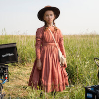 Young girl in a pioneer dress and hat standing in a grassy field on a film set, with camera equipment visible around her on a bright day.