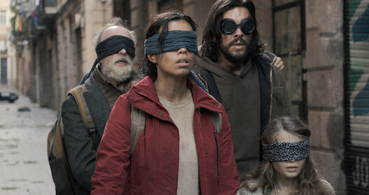 Three adults and a child walk through the street wearing blindfolds.