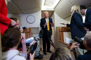 A man in a suit speaks to a group of reporters holding microphones in the aisle of a private jet, with cream-colored seats and overhead lighting.