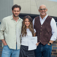 Three people standing outdoors, smiling at the camera. The woman in the middle holds a paper. They are dressed in casual and smart-casual attire, with a modern building structure visible in the background.