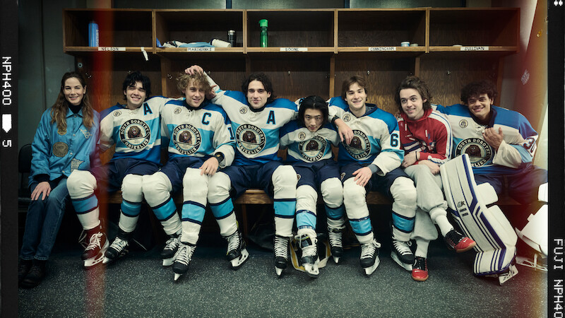 A group of young hockey players in blue and white uniforms and one in red sit together on a locker room bench, posing and smiling for a team photo, hockey gear and sticks by their sides.