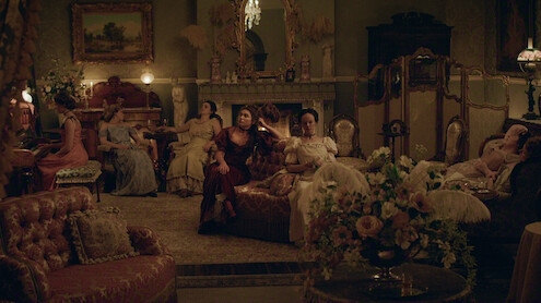 Women in elegant period dresses sit and lounge in a richly decorated parlor with vintage furniture, ornate decor, flowers, and a lit fireplace, creating a warm, historical atmosphere.