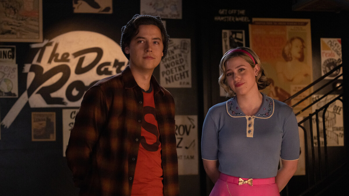 Cole Sprouse as Jughead Jones and Lili Reinhart as Betty Cooper stand together smiling in Season 7 of 'Riverdale.'