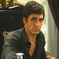 Man with serious expression sits at an ornate black chair with gold details, wearing a dark shirt and gold chain, indoors near a table in a formal setting with rich decor.