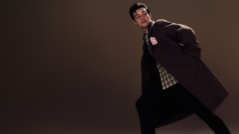 Louis Partridge stands on a dark brown background leaning over in an angled manner waring a brown coat with a plaid shirt and dark trousers.