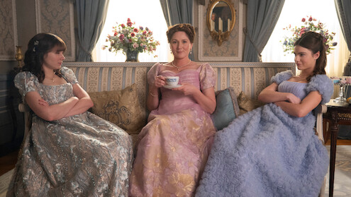 Three women in elegant period dresses sit on a vintage sofa in a richly decorated room, two looking displeased with arms crossed, one smiling with a teacup, creating a formal yet tense atmosphere.