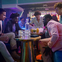 Five young men sit around a table in a lively nightclub, talking and laughing together. The table holds drinks, ice, and glasses, with colorful lighting and other people mingling in the background.