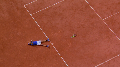 A tennis player lies on a clay court next to a tennis racquet, possibly celebrating or exhausted, viewed from above. The scene captures the lines and earthy texture of the tennis court.