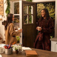 Two women smiling and talking in a cozy kitchen decorated for the holidays, holding mugs. Warm lighting, craft supplies on the counter, and greenery create a welcoming, festive atmosphere.
