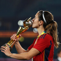 Female soccer player in red jersey kissing a gold trophy on a stadium field, wearing a medal and headband, celebrating victory with blurred stadium lights in the background.