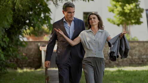 A man in a suit and a woman holding a jacket walk together outside on a sunlit path, surrounded by greenery and trees, with a building in the background.