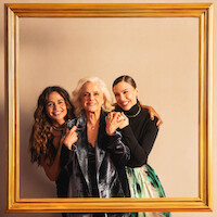 Um grupo de 9 atores da série 'Dom de Iludir' posa em um pequeno palco com o nome da série, sobre chão coberto por um tapete vermelho. As atrizes Nanda Costta, Marieta Severo e Alice Wegmann, no centro da foto, apoiam as mãos em uma grande moldura de quadro dourada